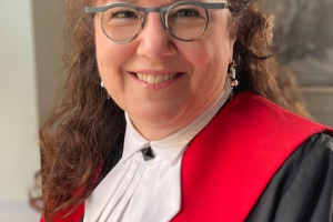 Woman with long curly brown hair and glasses in red, black and white judicial robes.