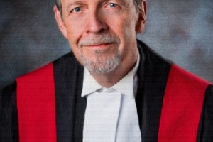 White man with a goatee wearing red, black and white judicial robes.